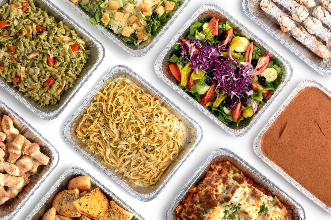 Large Italian catering spread with pasta trays, salad, garlic bread, and dessert